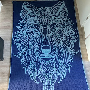May include: A blue and light blue crocheted blanket featuring a wolf design. The wolf's face is in the centre of the blanket and is surrounded by its fur.