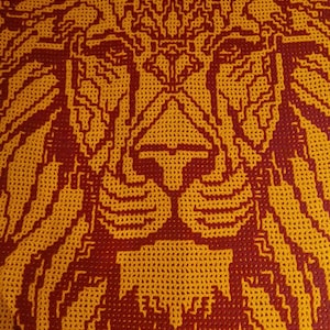 Puede incluir: Una manta de ganchillo con la cara de un león en hilo rojo y naranja. La cara del león está detallada con intrincados patrones y un diseño simétrico.
