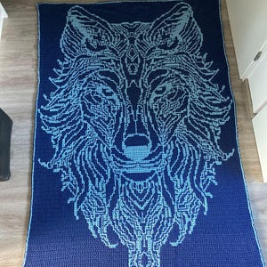 May include: A blue and light blue crocheted blanket featuring a wolf design. The wolf's face is in the centre of the blanket and is surrounded by its fur.