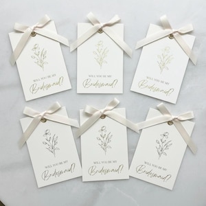 Puede incluir: Seis etiquetas rectangulares de papel blanco con un delicado dibujo floral en líneas doradas y el texto impreso Will You Be My Bridesmaid? Cada etiqueta tiene un ojal metálico dorado y un lazo de satén color crema atado.