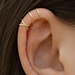 May include: Close-up of an ear adorned with gold-colored ear cuffs. The ear cuffs include three simple bands and a criss-cross design, creating a layered effect. The jewelry is set against the backdrop of brown hair.