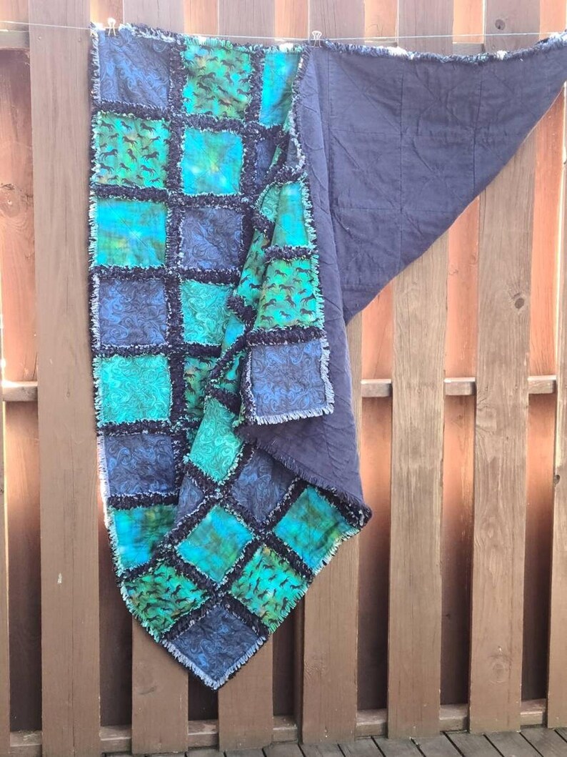 The quilt hangs vertically against a wooden fence with a folded corner revealing the solid dark blue backing fabric.