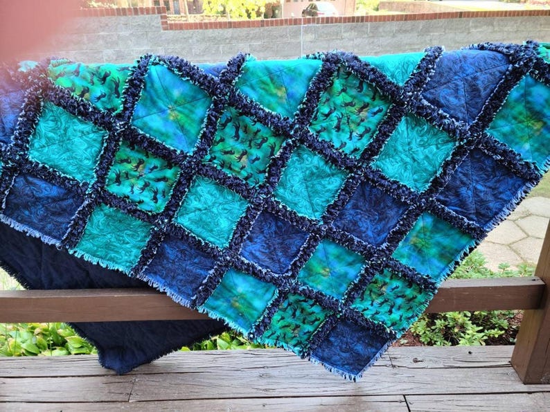 The rag quilt is draped over a wooden railing outdoors, showing its quilted squares and exposed fringed seams.