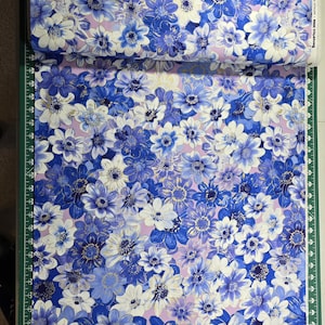 May include: A roll of fabric featuring a floral pattern with blue, white, and gold flowers on a light purple background. The fabric is on a green cutting mat.