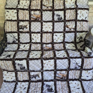 May include: A handmade patchwork quilt featuring a rustic woodland theme. The quilt showcases a grid pattern with alternating squares of plaid, animal prints, and textured brown fabric. The animal prints include deer, moose, and other wildlife.