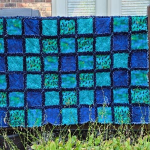 The full quilt is displayed flat against a brick wall and wooden railing, highlighting its alternating colored blocks.