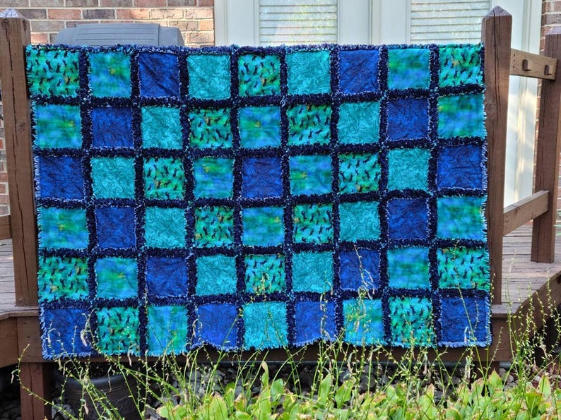 The full quilt is displayed flat against a brick wall and wooden railing, highlighting its alternating colored blocks.