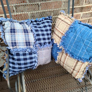 May include: Two square throw pillows with a patchwork design. The pillows feature a mix of blue and beige plaid fabrics, with a fringed edge. The pillows are placed on a metal chair, with a brick wall in the background.