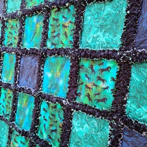 Close-up angled view of a rag quilt featuring squares of blue and teal marbled fabric alongside blocks with horse silhouettes.