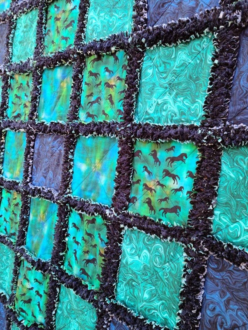 Close-up angled view of a rag quilt featuring squares of blue and teal marbled fabric alongside blocks with horse silhouettes.