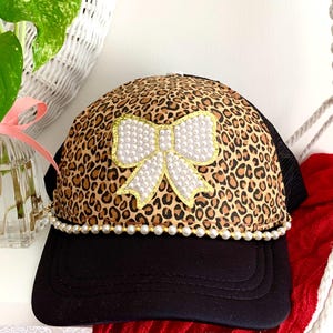 May include: A black and leopard print baseball cap with a pearl and gold trim. The cap features a large white bow embellished with pearls and gold trim. The cap is a stylish accessory.