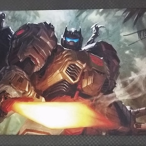 May include: A playmat with a detailed illustration of a Transformer character in a dynamic pose, with a glowing blade. The artwork showcases a combination of brown, gold, and red hues, set against a jungle backdrop. The Ultra Pro logo is visible.