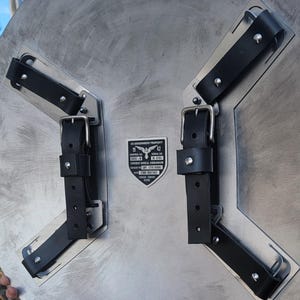 May include: A silver-colored shield with black leather straps and buckles. The shield has a textured surface and a metal plate with text that reads "US Government Property" and "Combat Shield Vibranium".