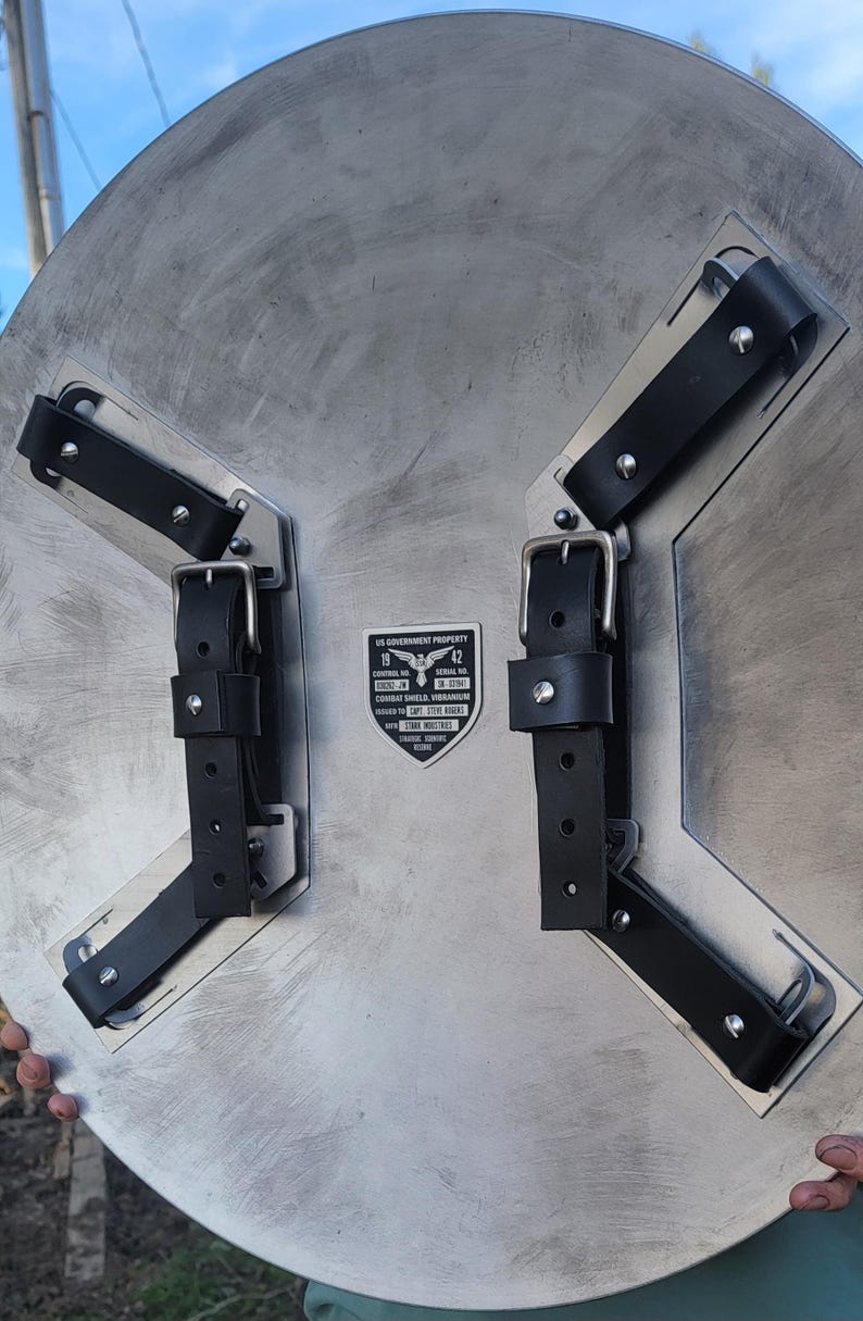 May include: A silver-colored shield with black leather straps and buckles. The shield has a textured surface and a metal plate with text that reads "US Government Property" and "Combat Shield Vibranium".