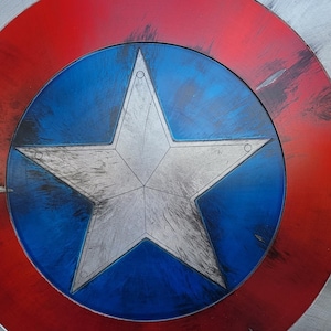 May include: A close-up of a Captain America shield. The shield features a red and silver outer ring, a blue center with a white star, and a distressed finish. The shield is a prop or collectible.