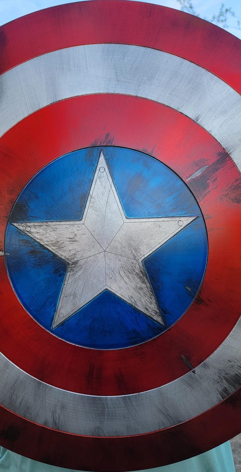 May include: A close-up of a Captain America shield. The shield features a red and silver outer ring, a blue center with a white star, and a distressed finish. The shield is a prop or collectible.