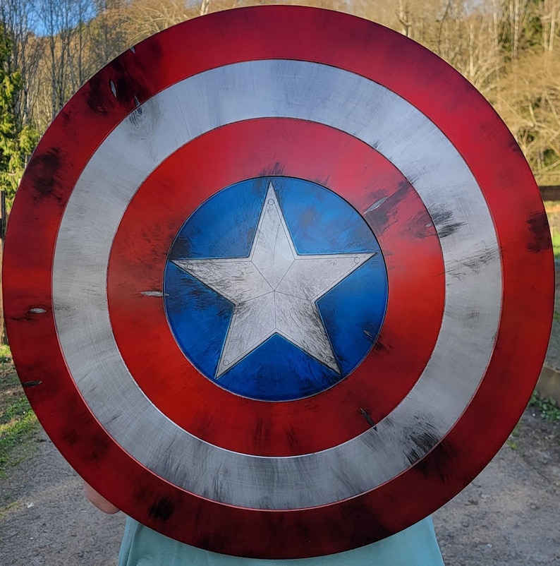 May include: A round shield with a distressed finish, featuring concentric rings of red, silver, and blue. A white star is centered on a blue circle. The shield is a replica of a superhero's iconic accessory.