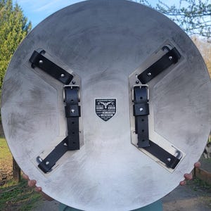 May include: A large, circular metal shield with a brushed silver finish. It features black leather straps and buckles on the back for securing the shield. The shield has a printed emblem with text that reads "US GOVERNMENT PROPERTY".