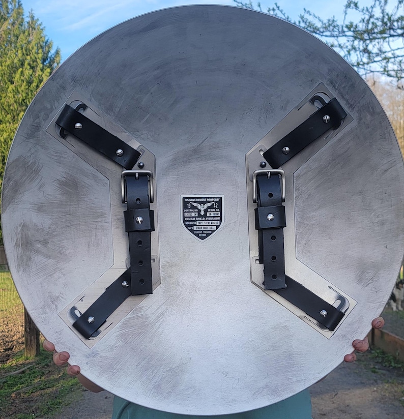 May include: A large, circular metal shield with a brushed silver finish. It features black leather straps and buckles on the back for securing the shield. The shield has a printed emblem with text that reads "US GOVERNMENT PROPERTY".
