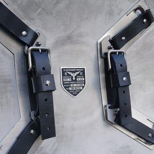May include: A silver-colored, circular shield with black leather straps and buckles. The shield has a brushed metal finish and a rectangular label with text that reads "US GOVERNMENT PROPERTY" and "COMBAT SHIELD, VIBRANIUM".