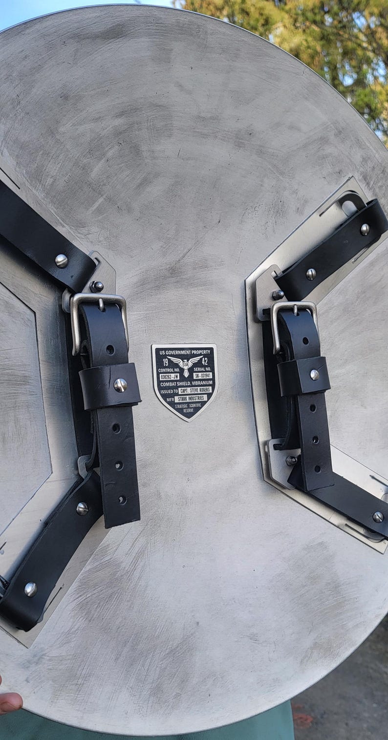 May include: A silver-colored, circular shield with black leather straps and buckles. The shield has a brushed metal finish and a rectangular label with text that reads "US GOVERNMENT PROPERTY" and "COMBAT SHIELD, VIBRANIUM".
