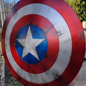 May include: A round shield with a red, white, and blue color scheme. The shield has a central blue circle with a white star, surrounded by white and red concentric rings. The shield has a distressed, weathered appearance.