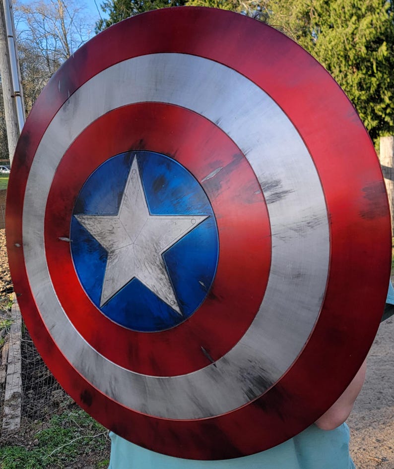 May include: A round shield with a red, white, and blue color scheme. The shield has a central blue circle with a white star, surrounded by white and red concentric rings. The shield has a distressed, weathered appearance.