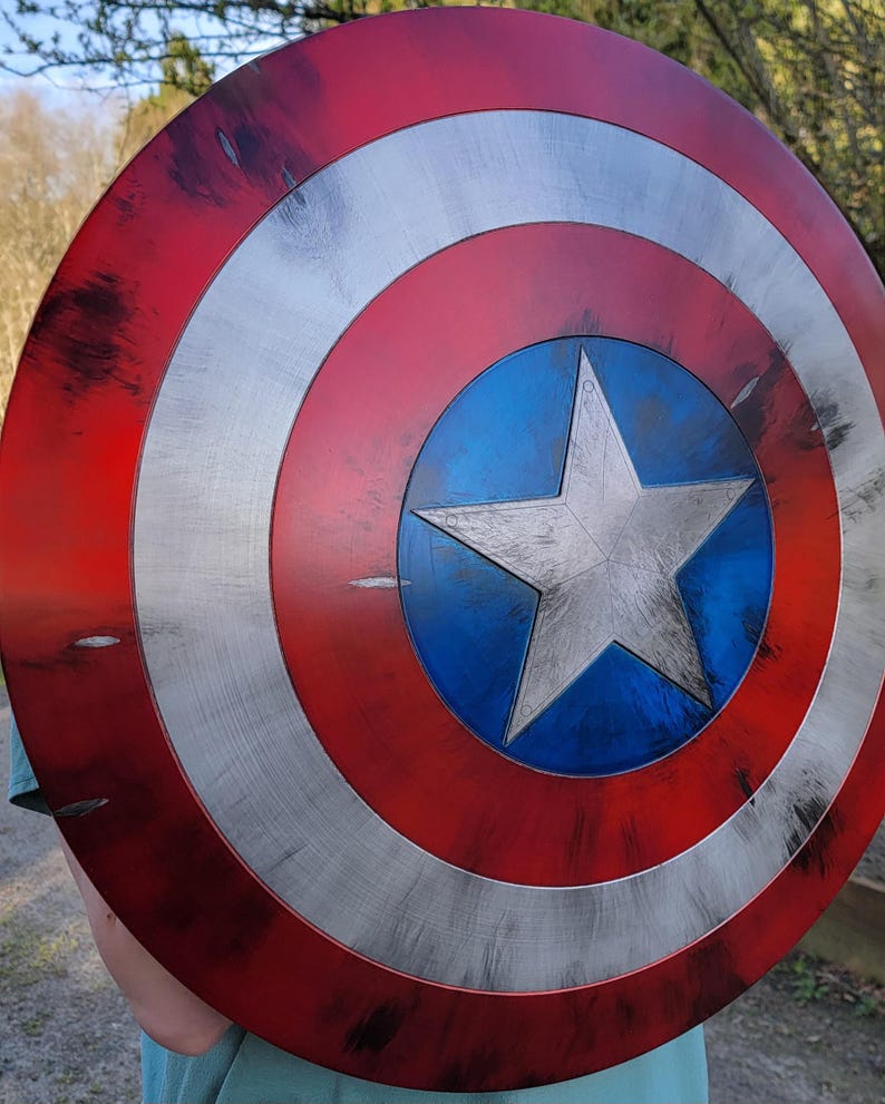 May include: A Captain America shield, featuring a red, white, and blue color scheme. The shield has a central blue circle with a white star, surrounded by concentric white and red bands. The shield has a distressed, battle-worn appearance.