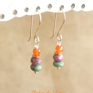May include: A pair of dangle earrings with silver hooks. Each earring features a stack of colorful beads: orange, gold, purple, and green. The earrings are suspended from a white, decorative element.