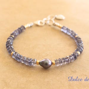 May include: A silver bracelet with a dark grey pearl and blue gemstones. The bracelet is made with a silver clasp and has a gold accent.