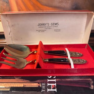 May include: A set of four gold-toned metal serving utensils with dark handles, presented in a red-lined box. The box lid features the text JOHNY'S GEMS, GUARANTEED QUALITY & PRICE (FACTORY), 199 SIKUK PHYA-SRI, BANGKOK, THAILAND, TEL. 21536.