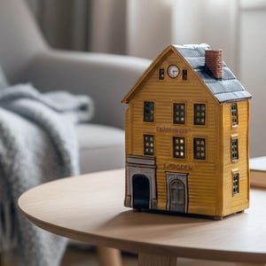 Puede incluir: Una casa decorativa amarilla con un reloj y varias ventanas está sobre una mesa de madera. La casa tiene un tejado gris y una pequeña chimenea. Una mano se extiende hacia una taza blanca sobre un libro.