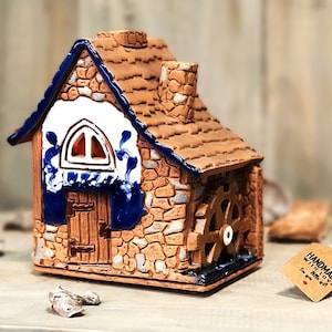 Könnte beinhalten: Ein kleines Keramikhaus mit braunem Dach, blauen Verzierungen und einem weißen Fenster. Das Haus hat ein kleines Wasserrad an der Seite und einen Schornstein oben. Das Haus steht auf einer Holzoberfläche mit einem braunen Etikett, auf dem "Handgefertigt mit Liebe für dich" steht.