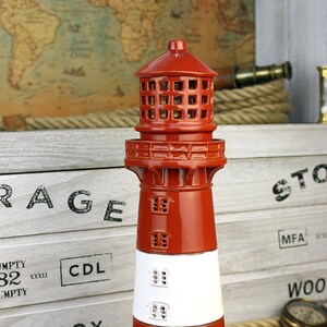 Op de afbeelding: Een rood en wit keramiek leuchtturmbeeldje met een gedetailleerd ontwerp. De leuchtturm heeft een klein wit gebouw aan de basis met een zwarte deur en ramen. De leuchtturm staat op een houten oppervlak.