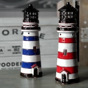 Può includere: Due figurine di faro in ceramica, una con strisce rosse e bianche e l'altra con strisce blu e bianche. Entrambe hanno tetti neri e piccole finestre.