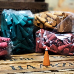 May include: Aromatic incense cones in various colors, including red, teal, and brown, are displayed in clear plastic bags. A single brown cone with a white tip stands in front of the bags on a wooden surface.