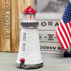 May include: A white lighthouse figurine with a red roof and a small red door. The lighthouse is sitting on a wooden table with a wooden box and an American flag in the background.