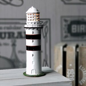 Op de afbeelding: Witte keramische vuurtorenfiguur met zwarte en rode strepen. De vuurtoren heeft een kleine groene basis en een licht in de bovenkant.