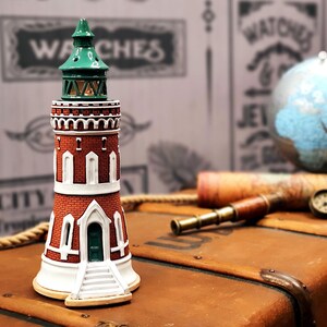 Op de afbeelding: Een miniatuur-lighthuisfiguur met een rode bakstenen basis, witte versieringen en een groen dak. De vuurtoren staat op een bruin houten oppervlak met een globe en een telescoop op de achtergrond.