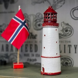 Op de afbeelding: Een wit en rood keramiek leuchttormodel met een rood dak en een klein raam. De leuchttorm staat op een houten oppervlak voor een Noorse vlag.