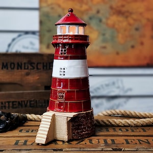 Puede incluir: Una figurita de faro de cerámica roja y blanca con una pequeña luz en su interior. El faro tiene una base blanca con escalones que conducen a la estructura principal. La figurita está sentada sobre una superficie de madera con una cuerda.