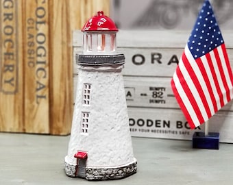 May include: A white lighthouse figurine with a red roof and a small red door. The lighthouse is sitting on a wooden table with a wooden box and an American flag in the background.