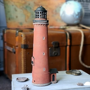 Puede incluir: Un faro en miniatura detallado con un exterior de ladrillo rojo, parte superior negra y pequeñas ventanas. El faro es un objeto decorativo, probablemente un objeto de colección de temática náutica. Se encuentra sobre una superficie blanca, con un fondo borroso de un globo terráqueo y una maleta vintage.