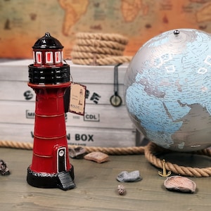 Op de afbeelding: Een rood en zwart vuurtorenbeeldje met een wit label waarop "Candle Lighthouse" en "Inslusive" staat. Het beeldje staat op een houten doos met een globe op de achtergrond.