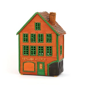 Könnte beinhalten: Ein kleines, orangefarbenes Keramikhaus mit grünen Fenstern und einem schwarzen Sockel. Das Haus hat ein Schild mit der Aufschrift "Bryggen Husflid".