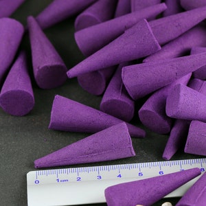 May include: A collection of purple incense cones, each with a conical shape and a flat base. A ruler with inch markings is placed near the cones. The cones are arranged on a dark surface.