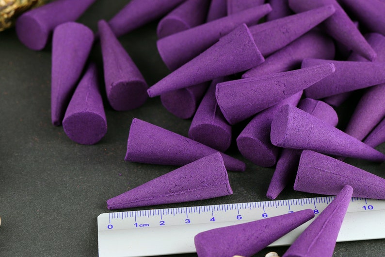 May include: A collection of purple incense cones, each with a conical shape and a flat base. A ruler with inch markings is placed near the cones. The cones are arranged on a dark surface.