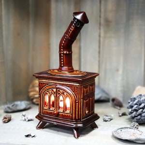 Pode incluir: Um aquecedor de fogão de cerâmica com acabamento marrom e dourado. O aquecedor tem uma chaminé e uma frente decorativa com uma janela que mostra uma luz brilhante no interior.