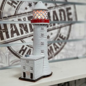 May include: White ceramic lighthouse figurine with a red roof and a light inside. The lighthouse has a small building attached to the base with the text "TUDOR ROCK LIGHTHOUSE 1835" on the side.