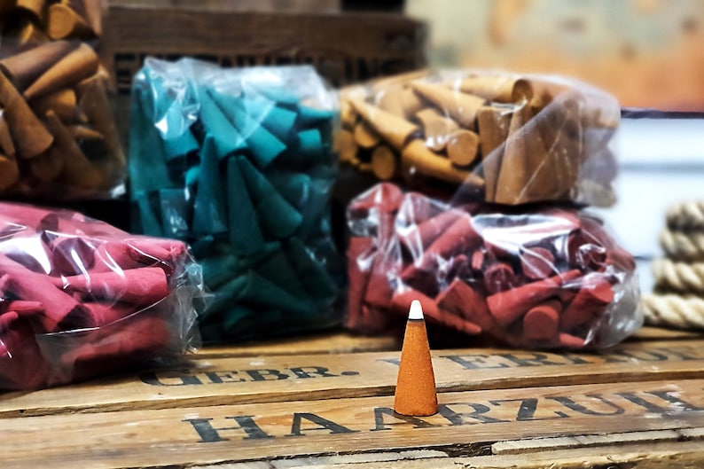 May include: Aromatic incense cones in various colors, including red, teal, and brown, are displayed in clear plastic bags. A single brown cone with a white tip stands in front of the bags on a wooden surface.